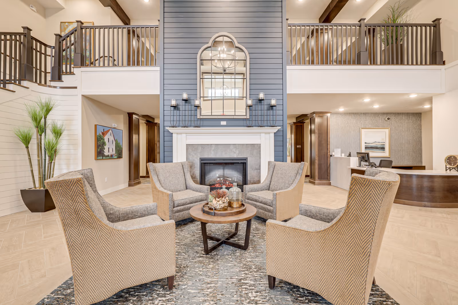 Lobby-style common area with four upholstered armchairs arranged around a round coffee table before a fireplace and an upper mezzanine balcony.