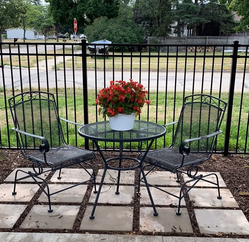 Outdoor patio area with two black metal chairs and a round black metal table with a white pot of red flowers on top, set on square stone tiles with a black metal fence and a street visible in the background.