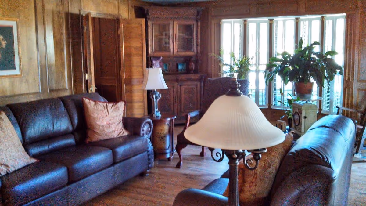 A cozy living room with dark leather sofas adorned with patterned cushions, a wooden side table with a lamp, a wooden cabinet, and large windows with white blinds letting in natural light. There are several green plants near the windows, and the room features wood-paneled walls and hardwood flooring.