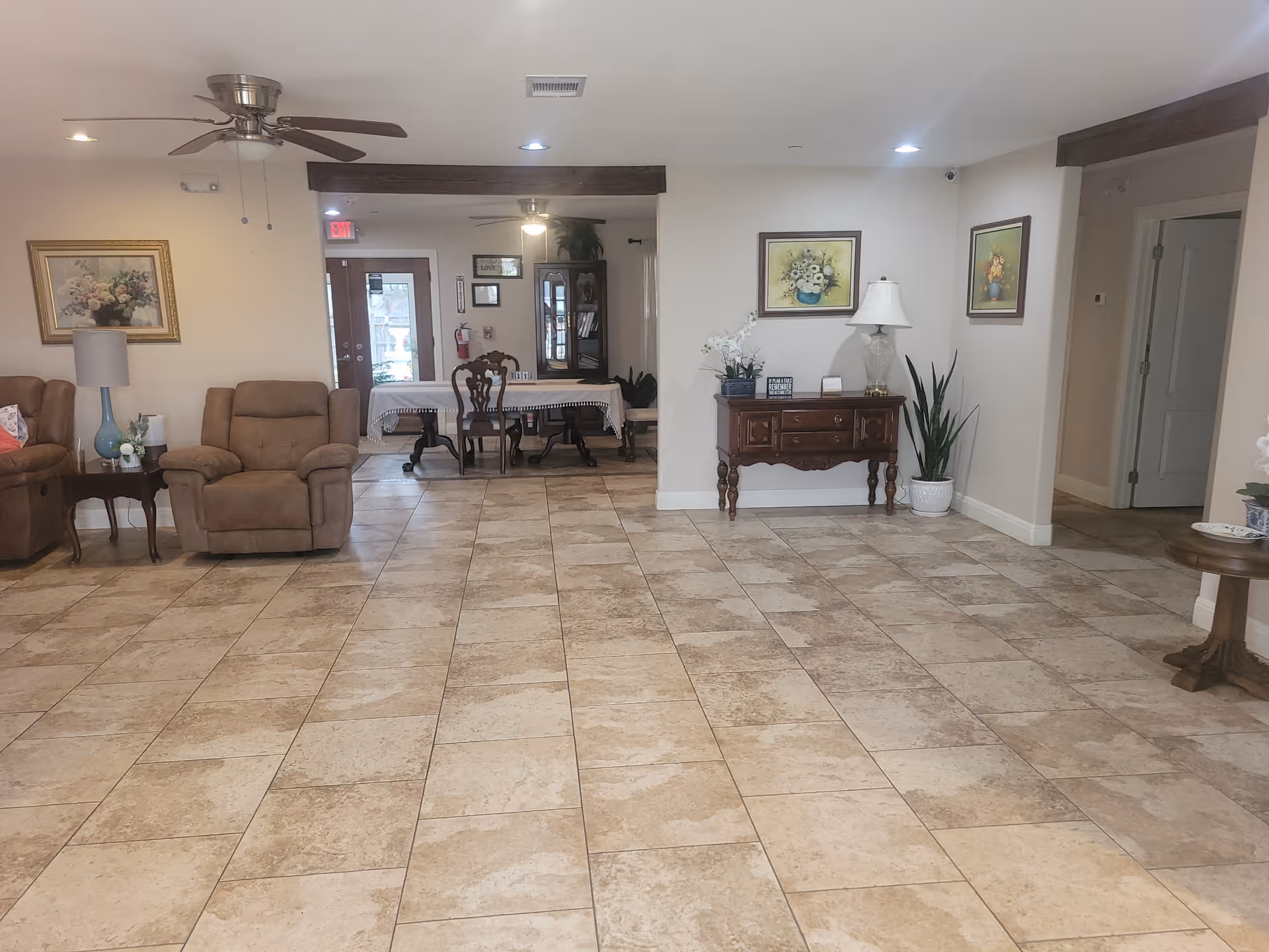 Spacious interior room with beige tiled floor, two brown recliner chairs on the left, a wooden side table with a blue lamp and flower vase, a wooden dining table with chairs in the background, and a wooden console table with a lamp and plants against the right wall. The walls are decorated with framed floral paintings, and ceiling fans with lights are visible.