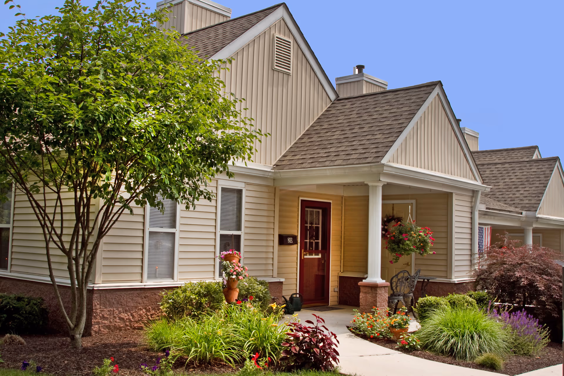 Front entrance of a beige single-story residential unit with a red door, porch column, and landscaped garden.