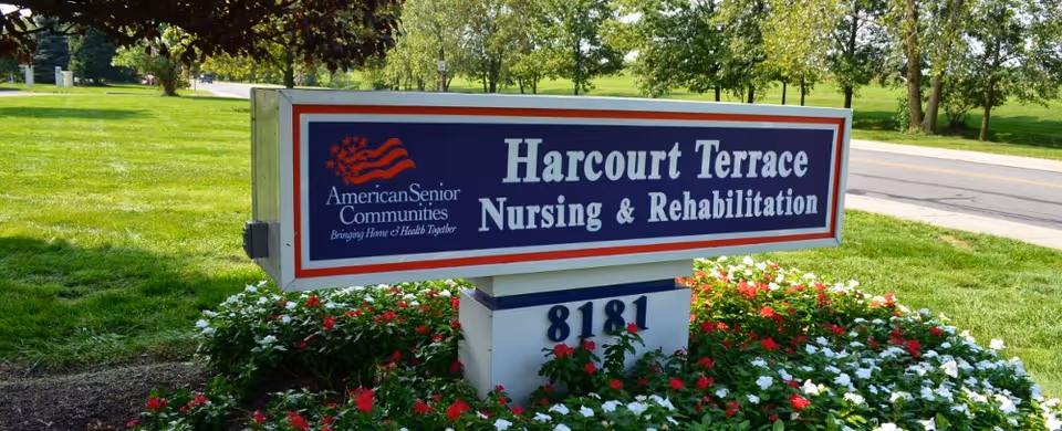 Outdoor sign for Harcourt Terrace Nursing & Rehabilitation surrounded by a flower bed with red and white flowers, green grass, and trees in the background near a road.