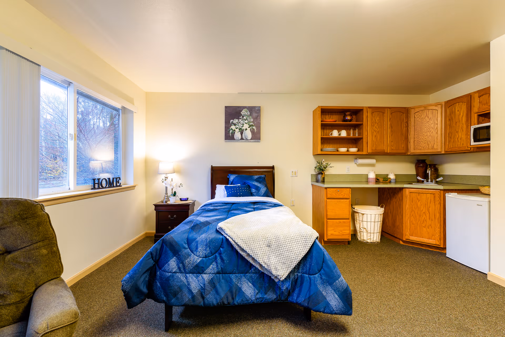 A cozy studio bedroom with a single bed, nightstand and lamp, a window and armchair, plus a small kitchenette with wooden cabinets.