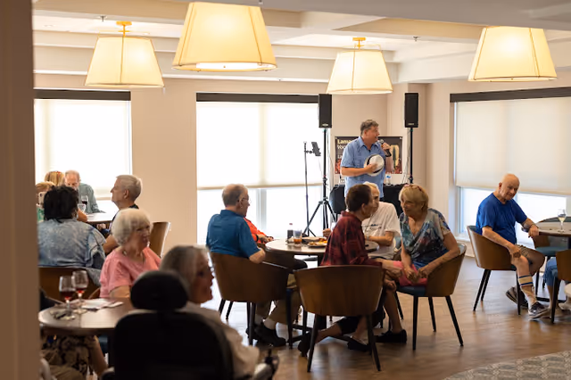 A group of older adults sit and chat at round tables in a bright common dining/activity room while a man performs at the front with a microphone.