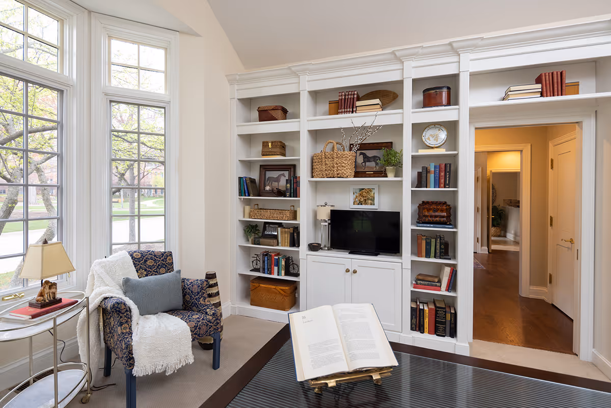 A cozy living room area with a large window letting in natural light. There is a patterned armchair with a white throw blanket and a gray pillow next to a small round side table with a lamp and books. A built-in white bookshelf filled with books, decorative baskets, and a small TV is against the wall. An open book is displayed on a stand on a dark coffee table in the foreground. A hallway with wooden flooring is visible through a doorway.