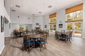 Bright communal dining room with several round wooden tables and chairs with blue cushions, large windows with woven shades, and wood-look flooring.