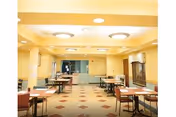 A well-lit communal dining room with multiple small tables and chairs arranged on a tiled floor under round ceiling lights.