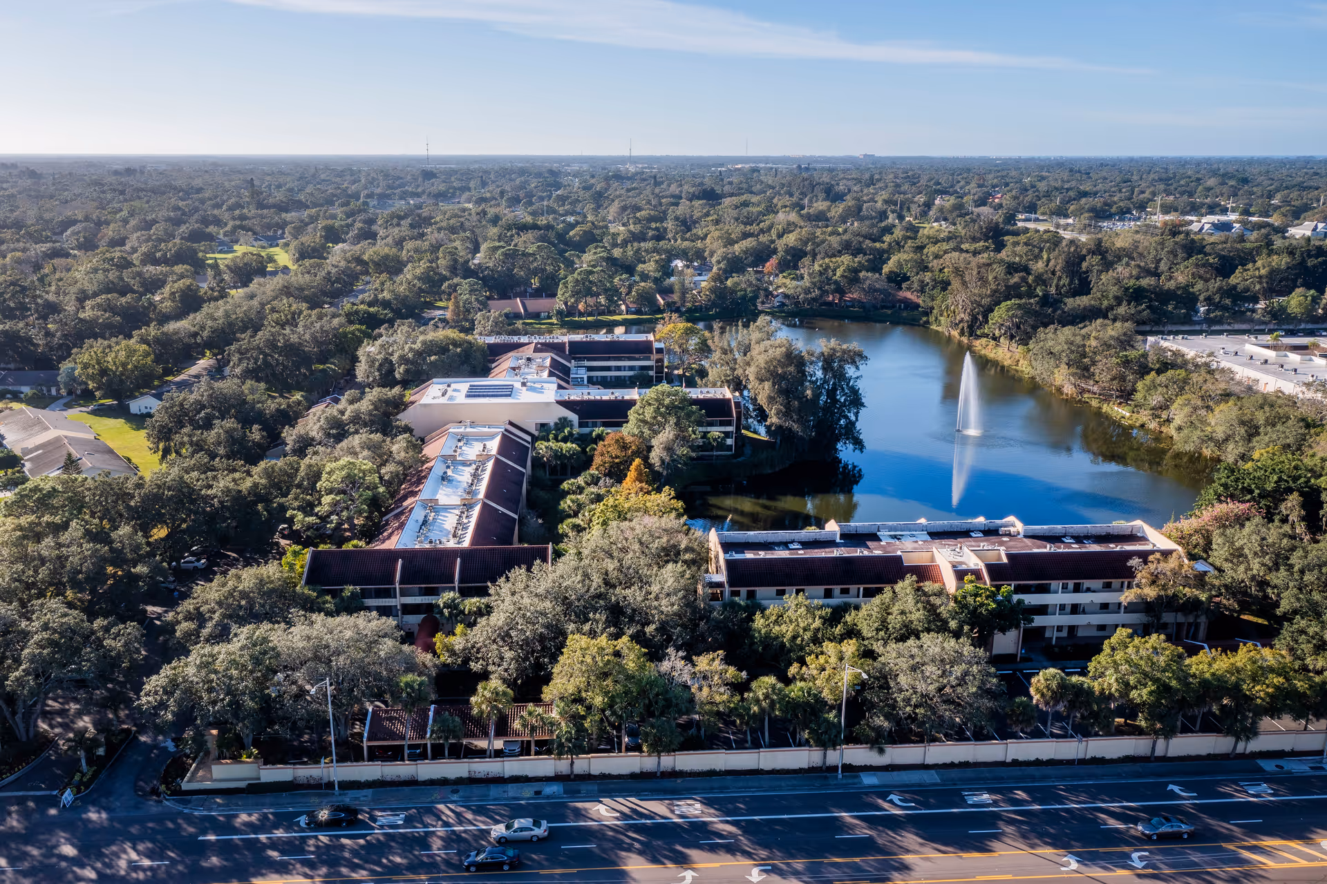 Aerial view of American House Sarasota senior living facility surrounded by trees, featuring multiple buildings with red roofs and a large pond with a fountain in the center. A road with cars is visible in the foreground.
