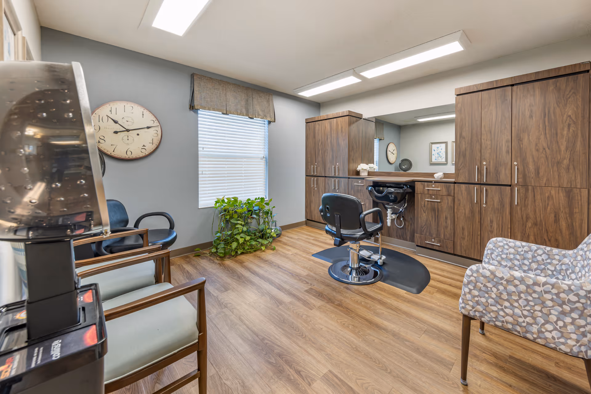 A well-lit salon-style room with a barber chair and sink, wood cabinets, seating, a wall clock and a potted plant.