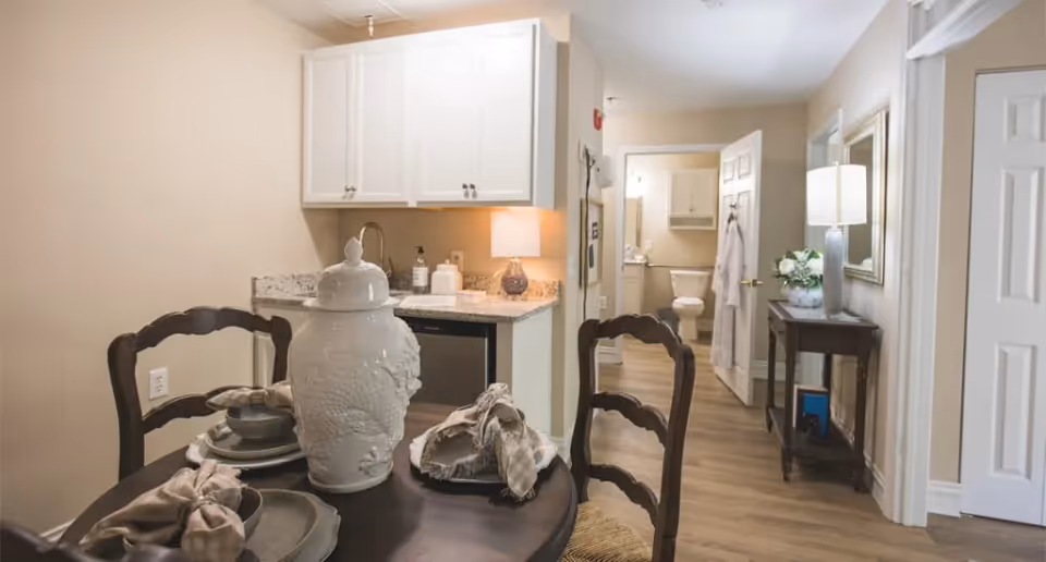 Small dining table with place settings and a decorative jar in a kitchenette area leading to a hallway with an open bathroom door.