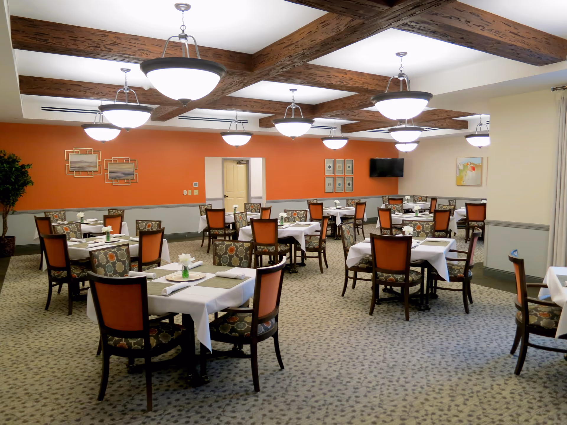 A dining room with multiple tables covered in white tablecloths, each set with placemats, napkins, and small flower vases. The chairs have patterned upholstery with orange backs. The room features a carpeted floor, orange and beige walls, wooden ceiling beams, and several hanging light fixtures. There is a flat-screen TV mounted on one wall and framed artwork on the walls.