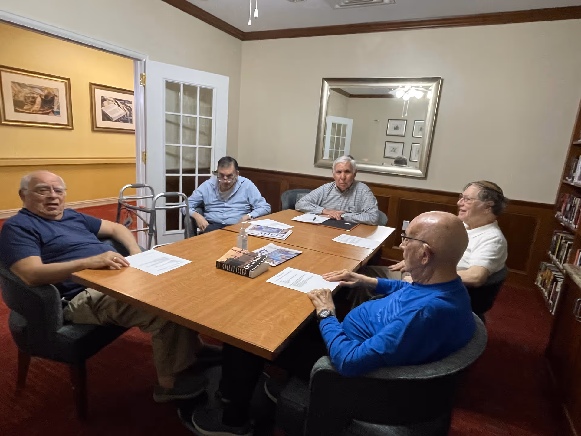 Five elderly men sitting around a wooden table in a room with beige walls and a large mirror. The table has papers, books, and a bottle of hand sanitizer on it. There is a walker near the door and bookshelves along one wall.