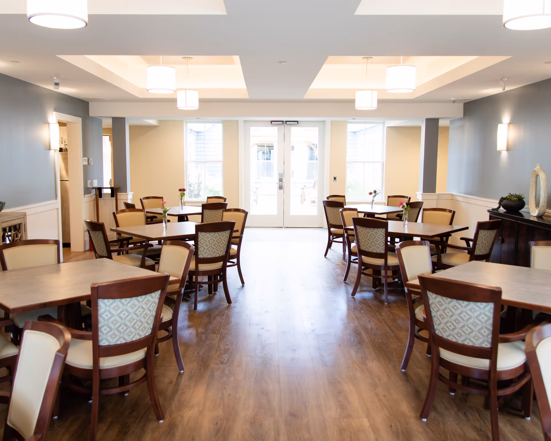 Bright communal dining room with multiple wooden tables and chairs, pendant lights, and double doors at the far end.