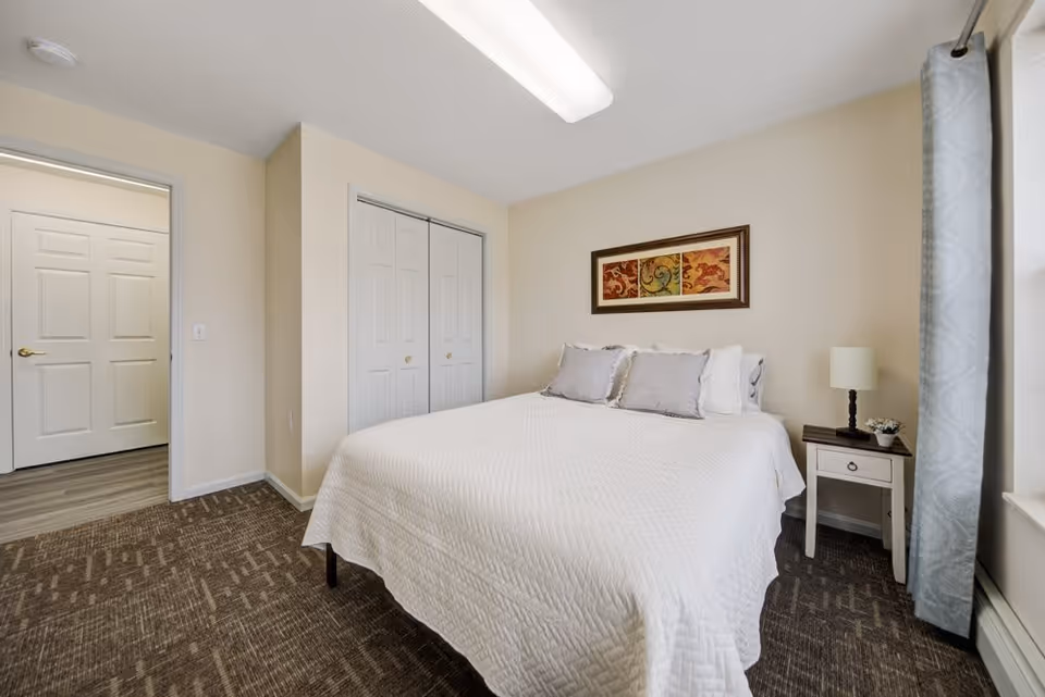 Neatly made bedroom with a bed, bedside table and lamp, framed artwork, closet doors and a window with curtains.
