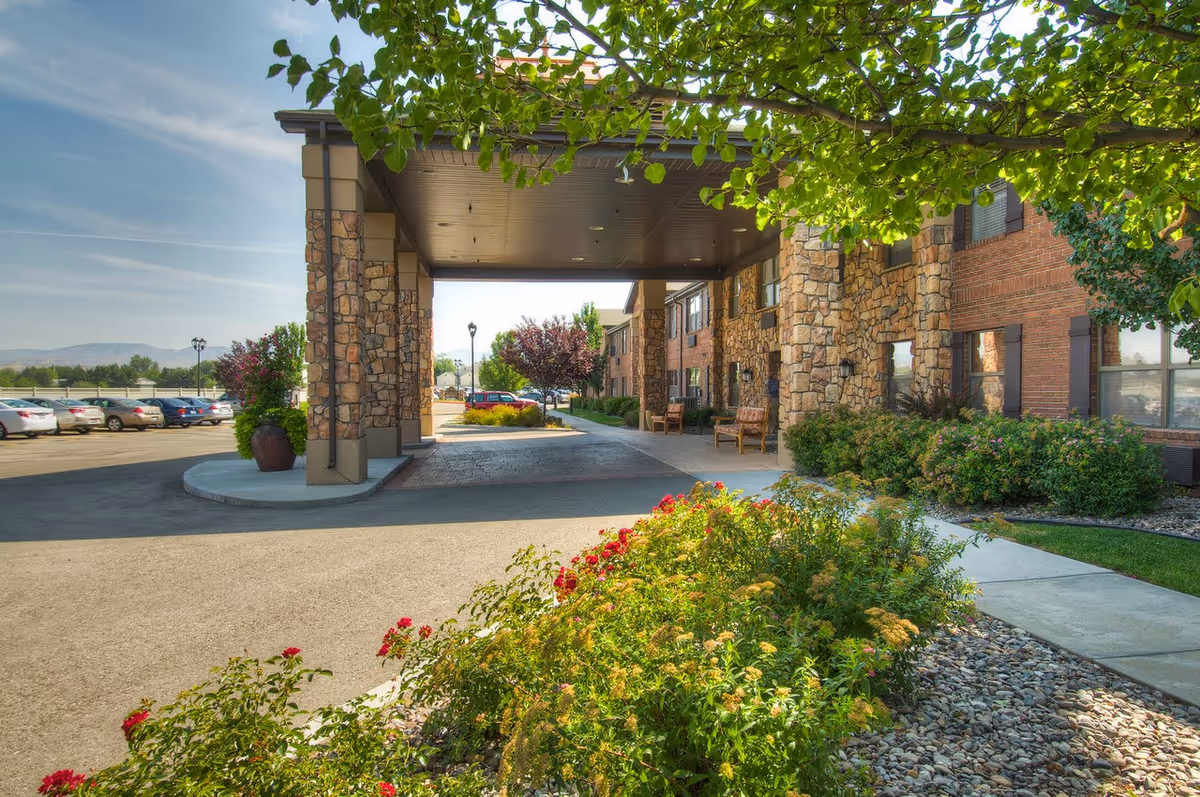 Entrance area of a senior living facility with a covered drop-off zone supported by stone pillars. There are benches along the building wall, landscaped bushes and flowers in the foreground, and a parking lot with cars in the background under a clear sky.