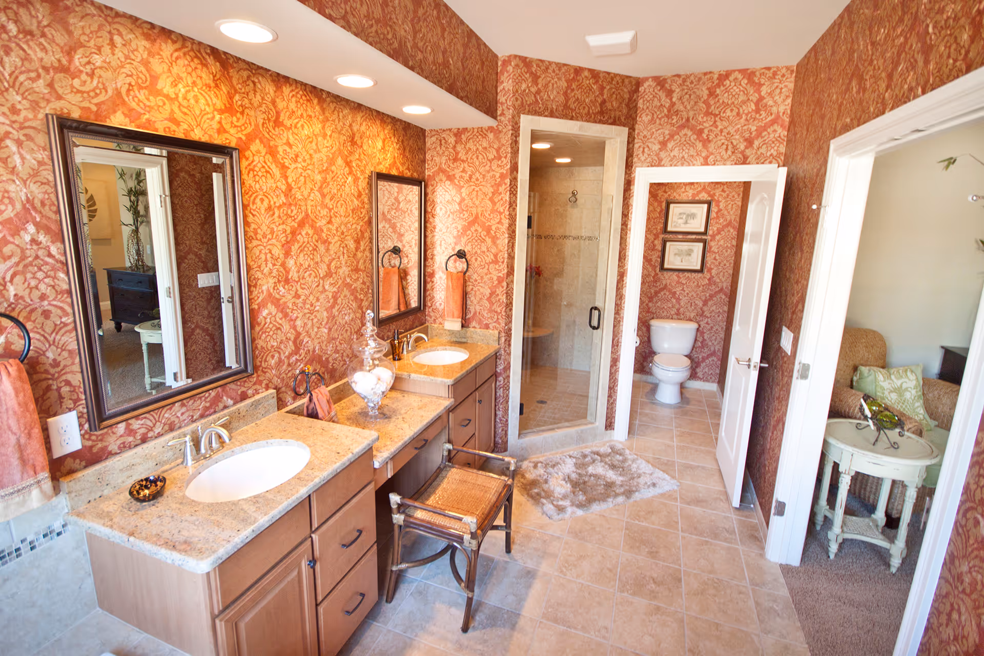 A spacious bathroom with two sinks set in granite countertops, wooden cabinets, and large mirrors above each sink. The walls are covered with red and gold patterned wallpaper. There is a glass-enclosed shower and a toilet in a separate alcove. A small wooden chair is placed in front of the sinks, and a plush rug is on the tiled floor. An adjacent room with a wicker chair and a small round table is visible through an open door.
