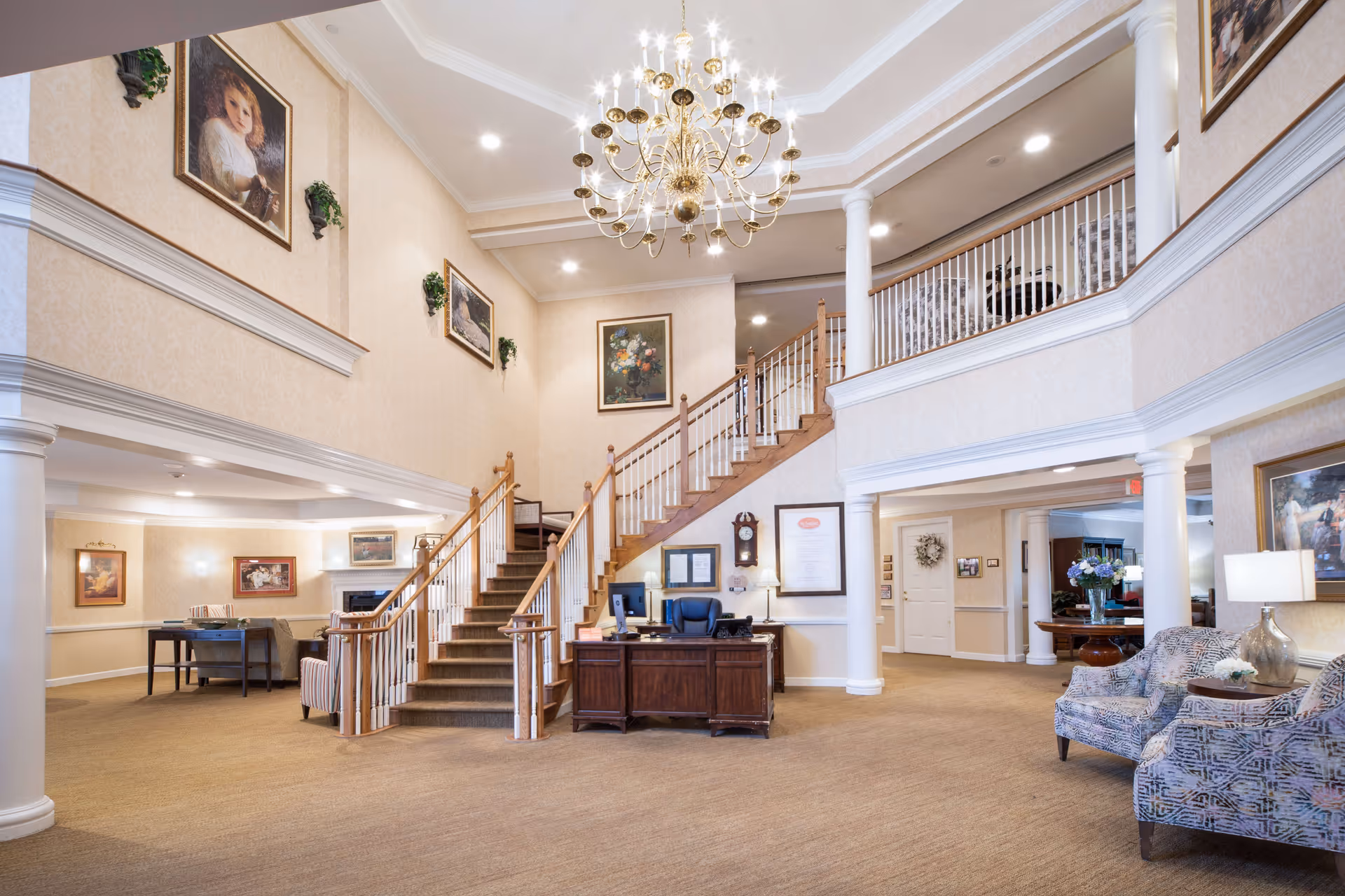 Spacious two-story senior living lobby with a central staircase, chandelier, reception desk, and seating areas.