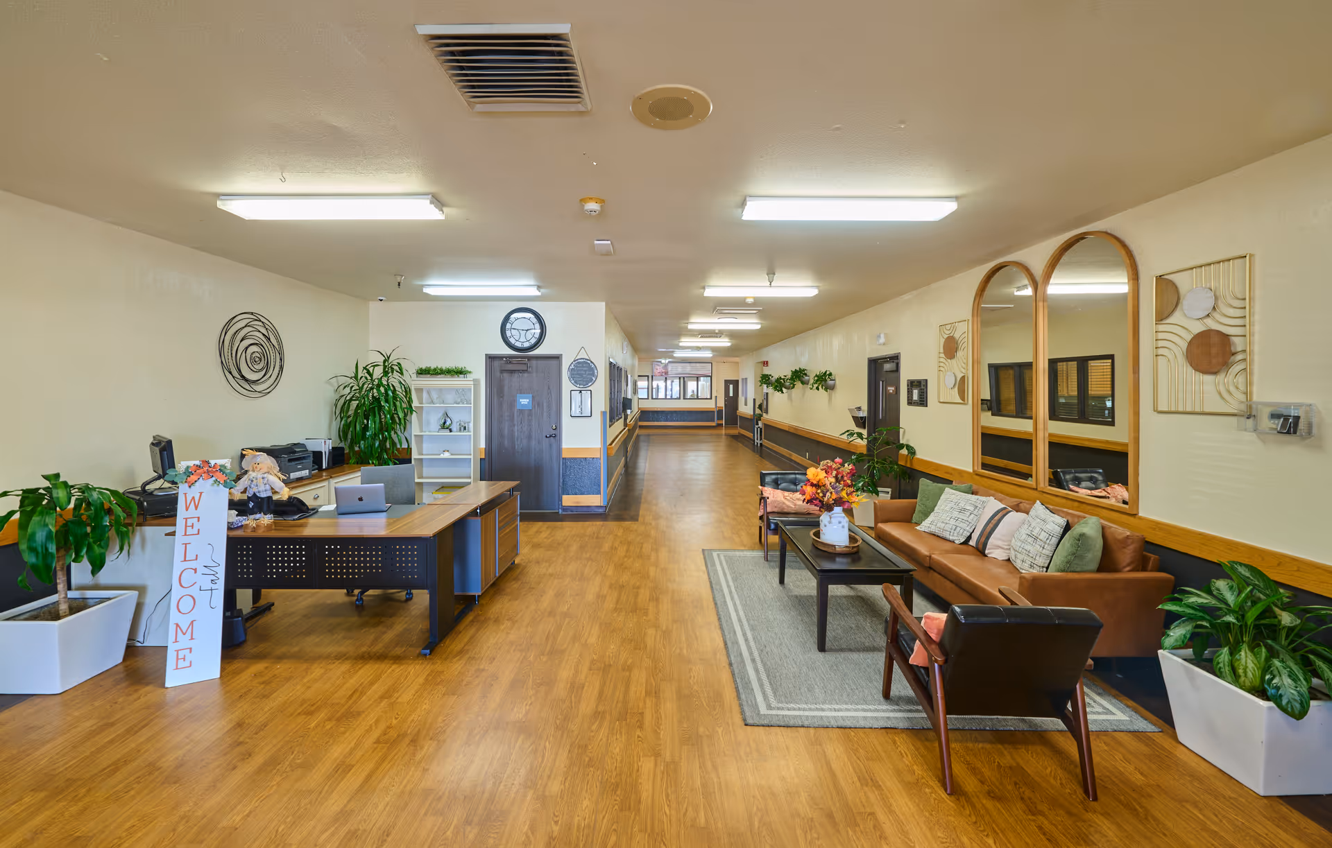 Interior view of a nursing and rehabilitation center hallway with a reception desk on the left, decorated with plants and a welcome sign. On the right, there is a seating area with a brown leather couch, a coffee table with a flower arrangement, and chairs. The walls are adorned with mirrors and artwork, and the floor is wooden.