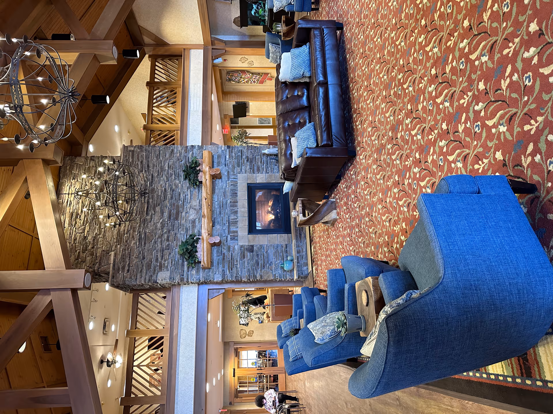 A spacious living room area with a high wooden ceiling and exposed beams. The room features a large stone fireplace with a wooden mantel, two wrought iron chandeliers hanging from the ceiling, a brown leather sofa with blue and white pillows, and several blue upholstered armchairs arranged around small wooden tables. The floor is covered with a red patterned carpet, and there is a balcony railing visible above the fireplace. In the background, a person is seated near a doorway, and there are plants and wall decorations throughout the space.