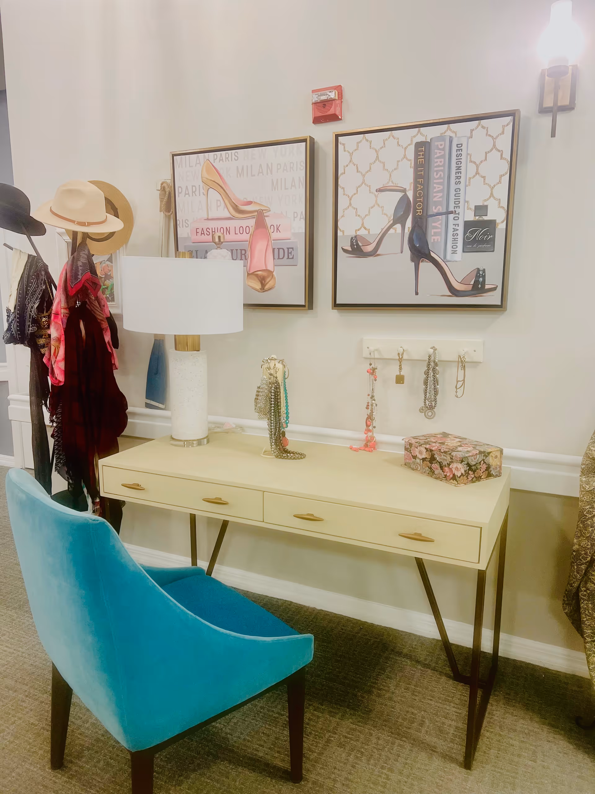 A stylish vanity area with a cream-colored desk featuring three drawers and a white lamp with a gold accent. A turquoise upholstered chair is placed in front of the desk. On the desk, there are several hanging necklaces and a floral decorative box. Two framed pictures of high-heeled shoes and fashion-related text hang on the wall above the desk. To the left, there is a coat rack with hats and scarves hanging on it. The room has beige walls and carpeted flooring.