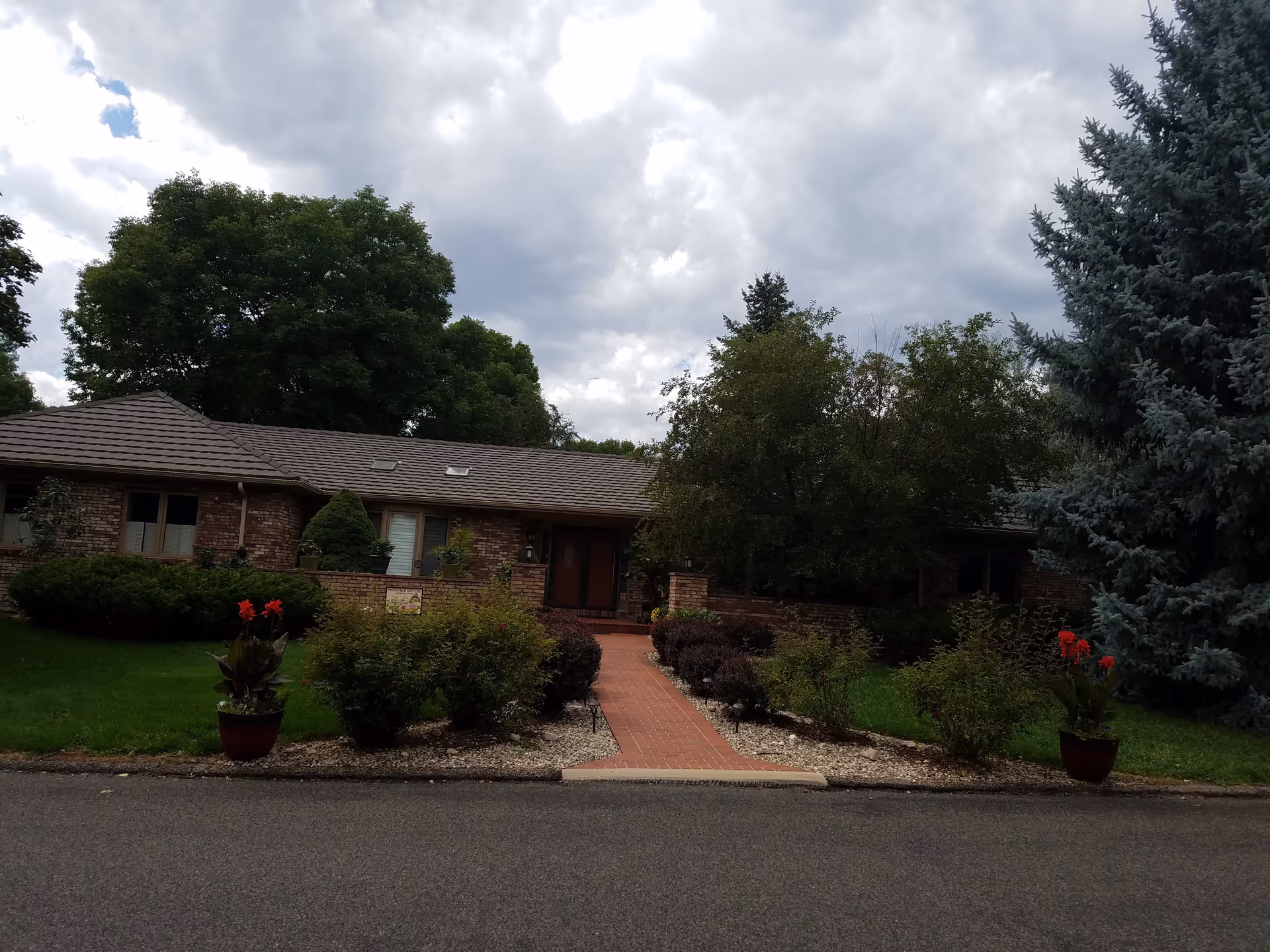 Front exterior of a single-story brick building with a central brick walkway, shrubs, potted flowers and large trees under a cloudy sky.