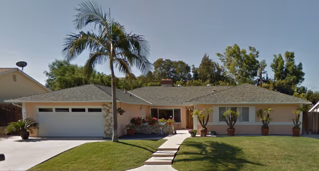 Single-story residential building with a gray shingled roof, beige exterior walls, and a two-car garage on the left. A concrete pathway leads from the driveway to the front door, flanked by potted plants and a palm tree in the front yard. The lawn is well-maintained with green grass and additional trees in the background under a clear blue sky.