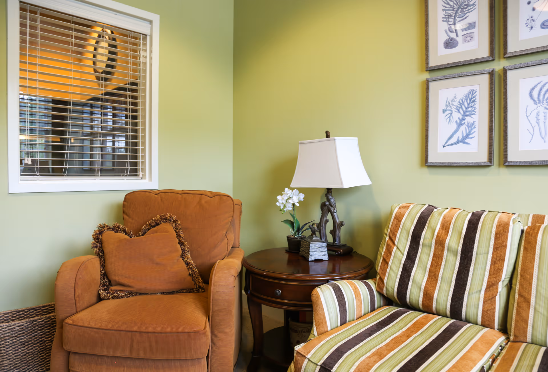 A cozy seating area with two upholstered chairs, a round side table holding a lamp and plant, and framed artwork on a green wall.