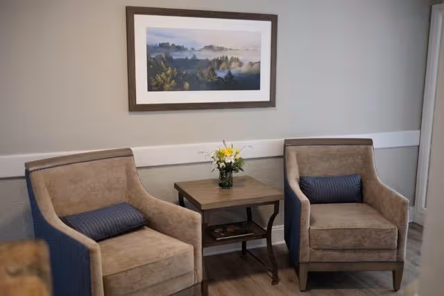 Two beige armchairs with blue accent pillows are positioned on either side of a small wooden side table with a vase of yellow and white flowers. A framed landscape photograph hangs on the light gray wall behind the chairs.