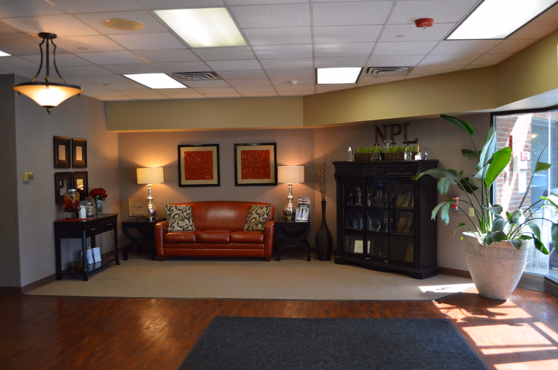 A cozy interior lounge area with a red leather couch adorned with two patterned pillows, flanked by two side tables each holding a lamp. Above the couch are two framed red artworks. To the right, there is a black cabinet with glass doors displaying various items and a large potted plant next to a window letting in natural light. On the left side, there is a small table with a beverage dispenser and flowers. The floor is a combination of wood and carpet, and the ceiling has recessed lighting and a hanging light fixture.