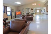 A spacious nursing home common area with leather armchairs and sofas arranged around a television. The room has large windows with curtains, a lamp on a side table, and a long hallway leading to other parts of the facility.