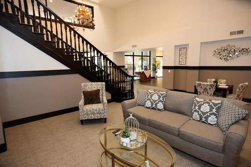 A spacious living room area in a senior living facility featuring a gray sofa with patterned cushions, a patterned armchair with a brown pillow, a glass coffee table with decorative items, and a black wooden staircase with a chandelier above. In the background, there is a dining area with a table and chairs, wall art, and large windows letting in natural light.