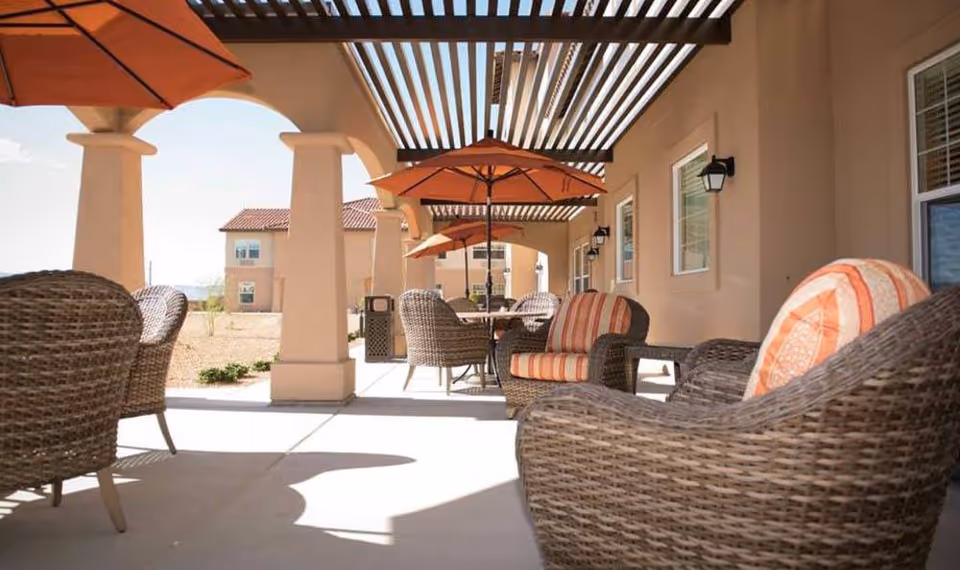 Outdoor patio area with wicker chairs and tables under a pergola. Orange umbrellas provide shade, and the building exterior is visible with windows and wall-mounted lights.
