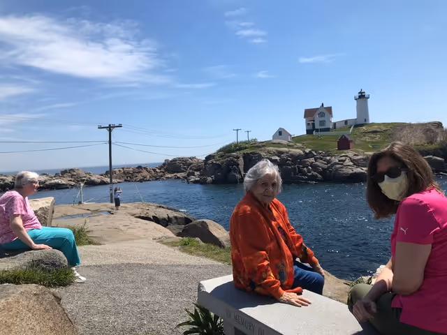 Three women sitting near a rocky waterfront with a lighthouse and small buildings on a hill in the background under a partly cloudy sky. One woman is wearing a pink shirt and a face mask, another is in an orange jacket, and the third is in a pink top and teal pants.