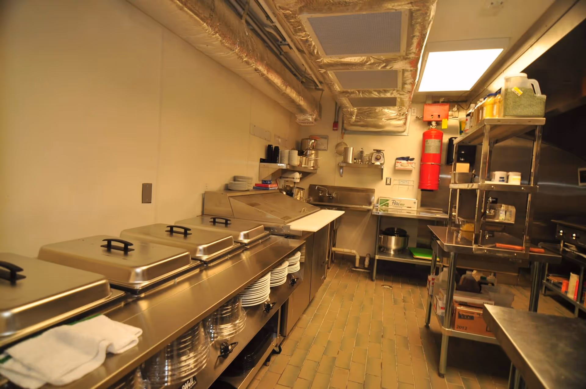 Commercial stainless-steel kitchen with food warmers, shelving, sinks and overhead ventilation ducts.
