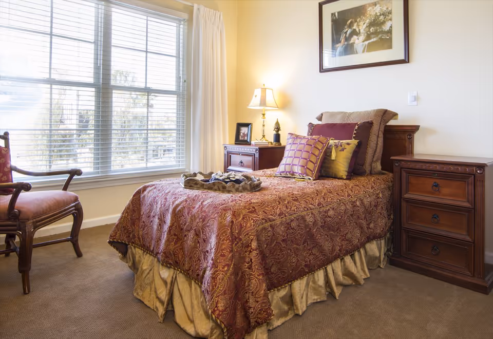 Bright, furnished bedroom with a single bed covered in patterned bedding, two wooden nightstands, a lamp, a framed picture, a window with blinds, and an upholstered chair.