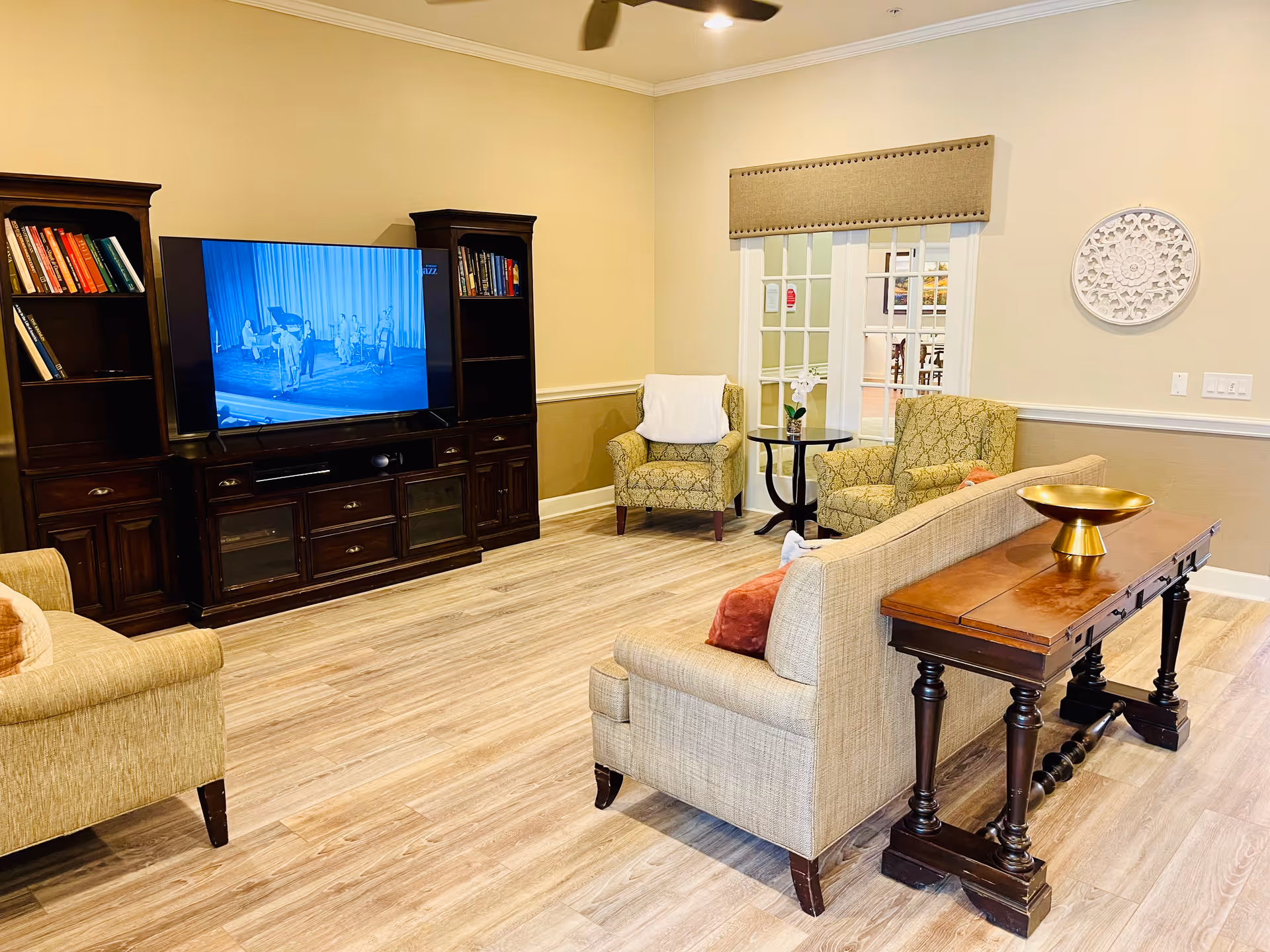 Well-lit common lounge with sofas, patterned armchairs, a large TV in a dark wood entertainment center, and a wooden console table.
