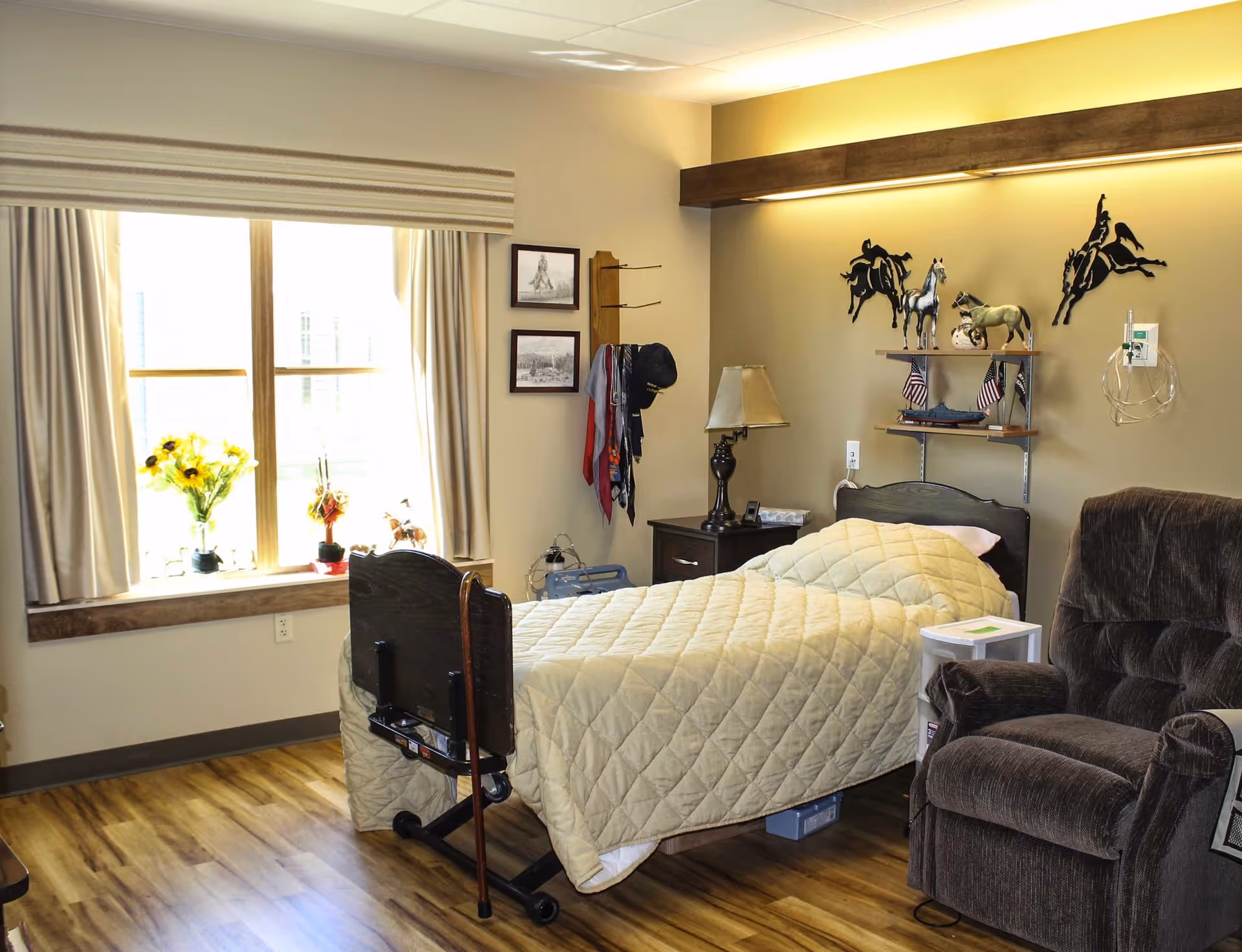 A cozy bedroom in a senior living facility featuring a single bed with a beige quilted blanket, a wooden headboard, and a bedside table with a lamp. There is a large window with beige curtains and a windowsill decorated with flowers. On the wall above the bed are decorative horse figurines and framed pictures. A comfortable brown recliner chair is positioned next to the bed, and the room has wood flooring.