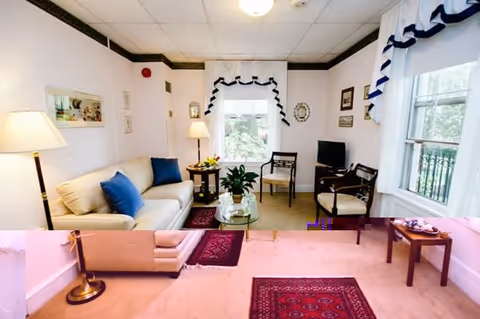 A cozy living room with a white sofa adorned with blue cushions, two chairs, a glass coffee table with a plant, a floor lamp, and a small TV on a stand. The room has white walls with dark trim, two windows with white curtains featuring blue accents, and red patterned rugs on a light carpeted floor.