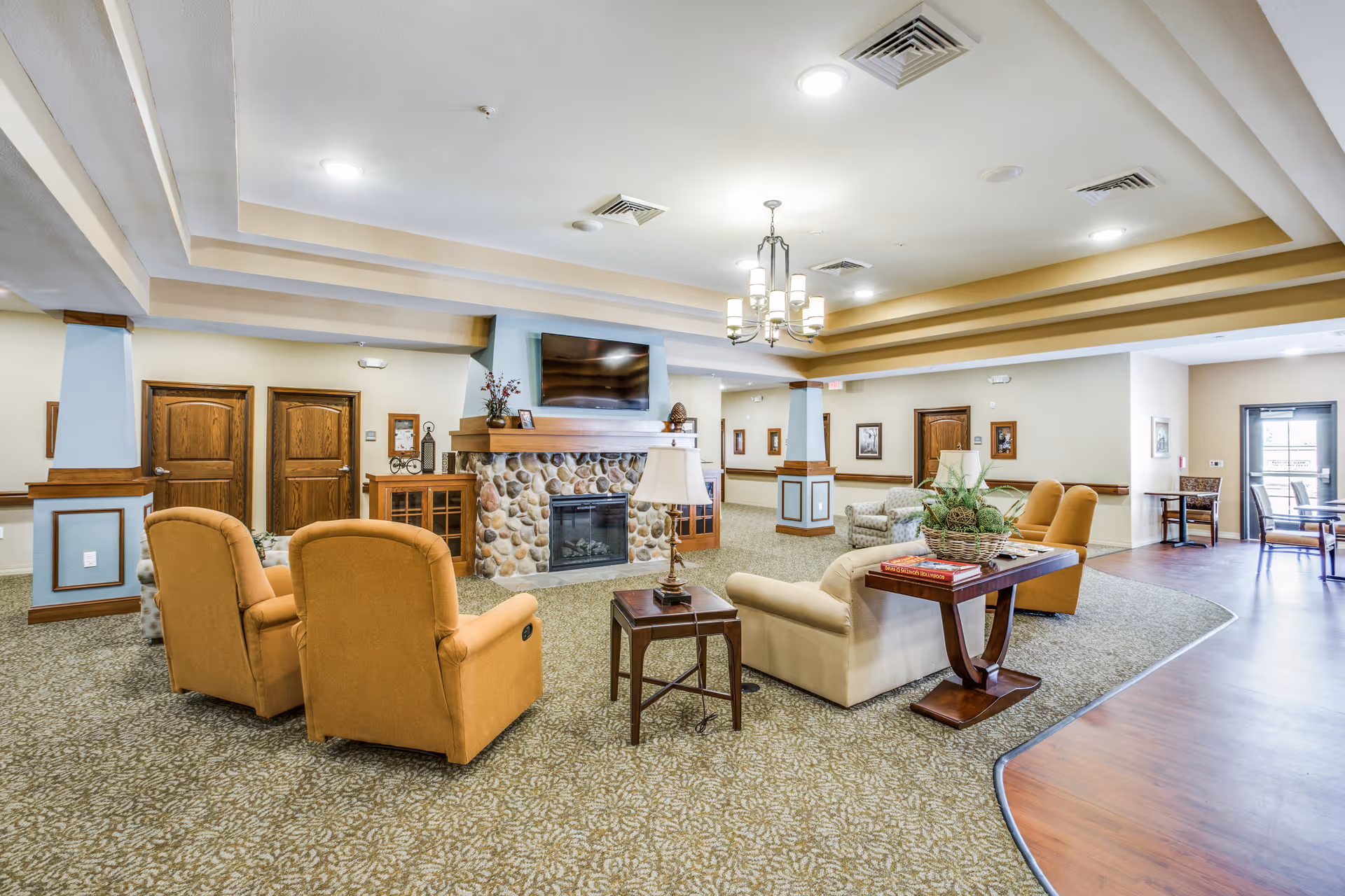 A spacious and well-lit living room area in a senior living facility featuring a stone fireplace with a mounted flat-screen TV above it. The room has comfortable seating including two mustard yellow armchairs, a beige sofa, and a patterned armchair. There are side tables with lamps and decorative items, a chandelier hanging from the ceiling, and framed pictures on the walls. The floor is carpeted with a patterned design, and there is a wooden floor section near the entrance door with additional seating and a small table.