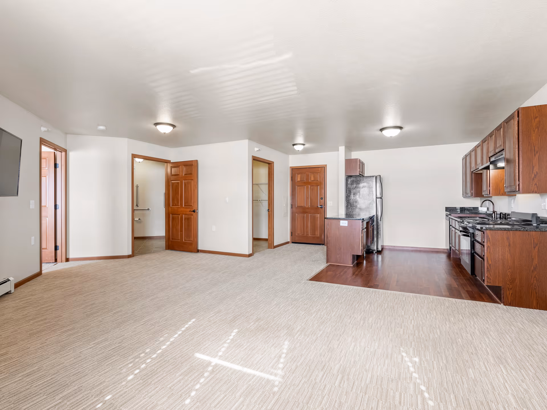 An empty senior living apartment interior featuring a combined living room and kitchen area. The living room has beige carpet flooring and a wall-mounted TV. The kitchen has dark wood cabinets, black granite countertops, stainless steel appliances, and hardwood flooring. There are three wooden doors visible, one leading to a bathroom with grab bars and another to a closet. The walls are painted white and the ceiling has recessed lighting fixtures.