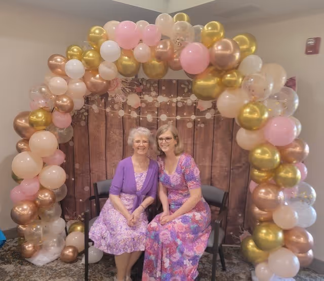 Two women sitting on chairs under a decorative balloon arch with pink, gold, white, and rose gold balloons. They are smiling and wearing floral dresses, with a wooden backdrop behind them.
