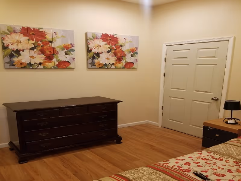 A bedroom with wooden flooring, a dark wooden dresser against a beige wall, two floral paintings above the dresser, a closed white door, and a bedside table with a black lamp and some items on it. Part of a bed with a floral and striped bedspread is visible in the foreground.