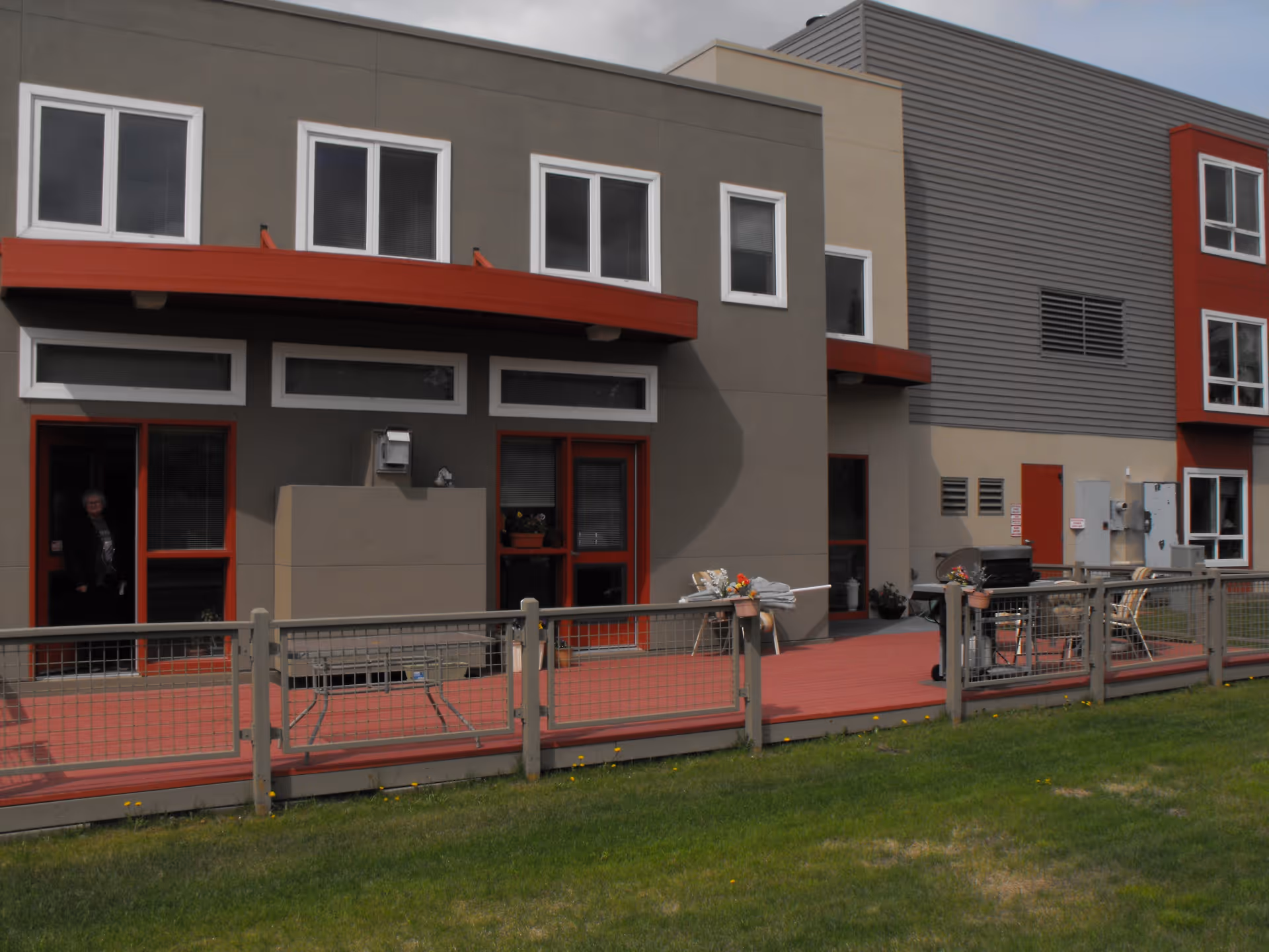 Exterior view of a modern senior living building with a red deck, patio furniture, and fenced lawn.