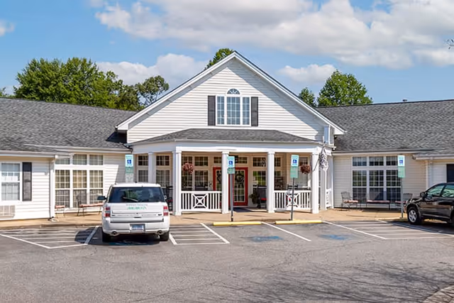 Front exterior view of a single-story senior living facility building with a peaked roof, white siding, and large windows. There are two cars parked in front in designated parking spaces, including handicap spots. The entrance has a covered porch with white columns and hanging flower baskets. Trees and a partly cloudy sky are visible in the background.