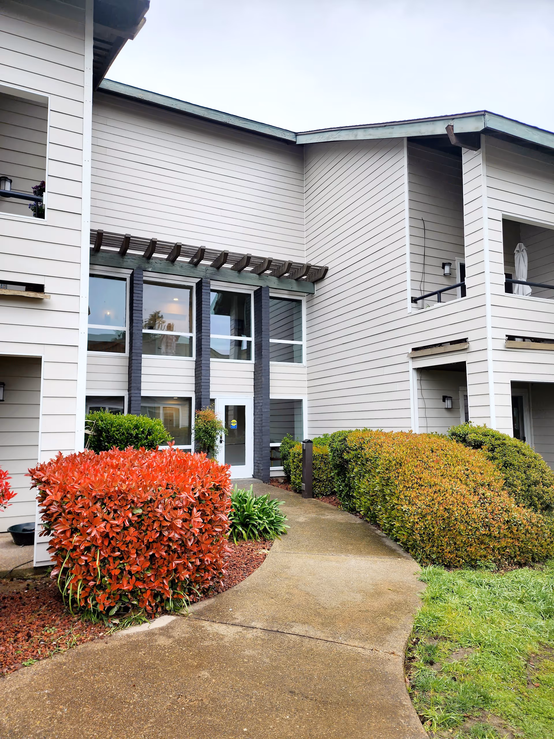 Exterior view of a two-story building with beige siding and black trim around the windows. There is a concrete walkway leading to a glass door entrance, surrounded by well-maintained bushes with red and green foliage. The sky is overcast.