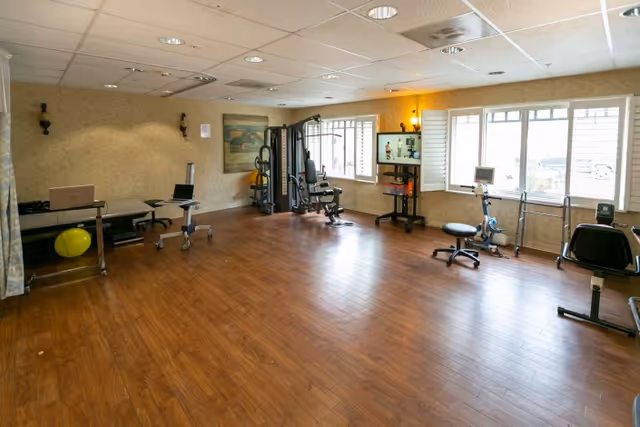 A spacious room with wooden flooring and beige walls, equipped with various exercise and physical therapy equipment including a multi-station gym machine, stationary bikes, a treatment table, and stools. The room has large windows allowing natural light to enter and a painting on the wall.