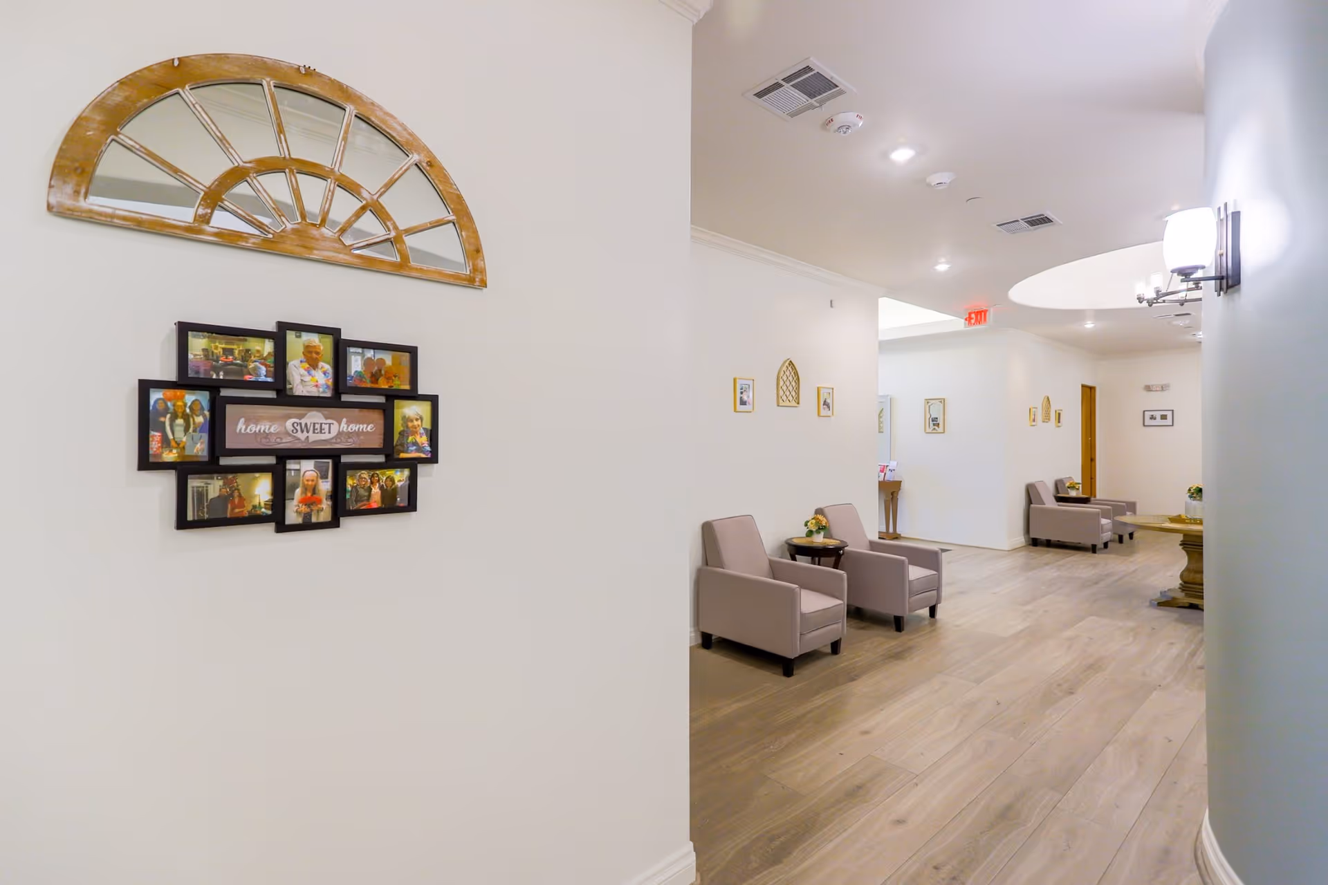 Bright memory care facility hallway with seating, a photo collage that reads "home sweet home," and a decorative half-moon mirror on the wall.