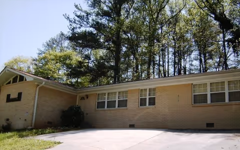 Exterior view of a single-story brick building with multiple windows, surrounded by tall trees and a clear blue sky. There is a concrete driveway in front of the building.