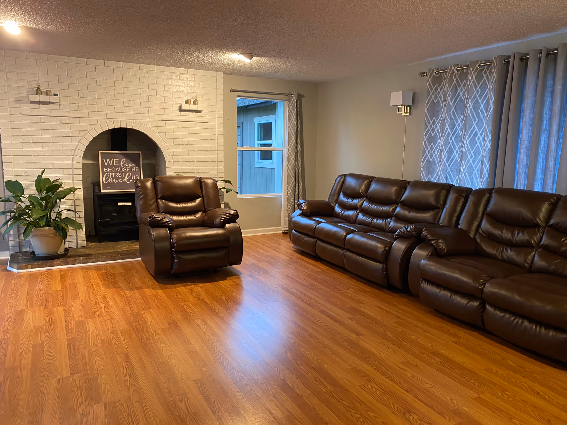 A living room with wooden flooring, featuring a large brown leather sofa and a matching recliner chair. There is a white brick fireplace with a black stove insert and a decorative sign that reads 'We love because He first loved us.' A potted plant is placed to the left of the fireplace. The room has two windows with patterned curtains and a wall-mounted light fixture.