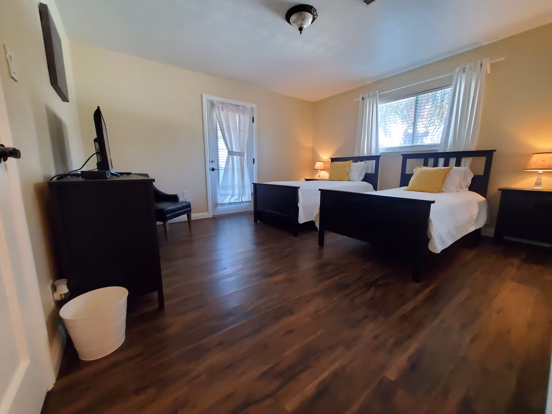 A bedroom with two single beds featuring dark wooden frames, white bedding, and yellow accent pillows. There is a window with white curtains above the beds, two bedside tables with lamps, a dark wooden dresser with a TV on top, a black chair, and a white waste bin on the wooden floor. A door with a sheer curtain is visible in the background.